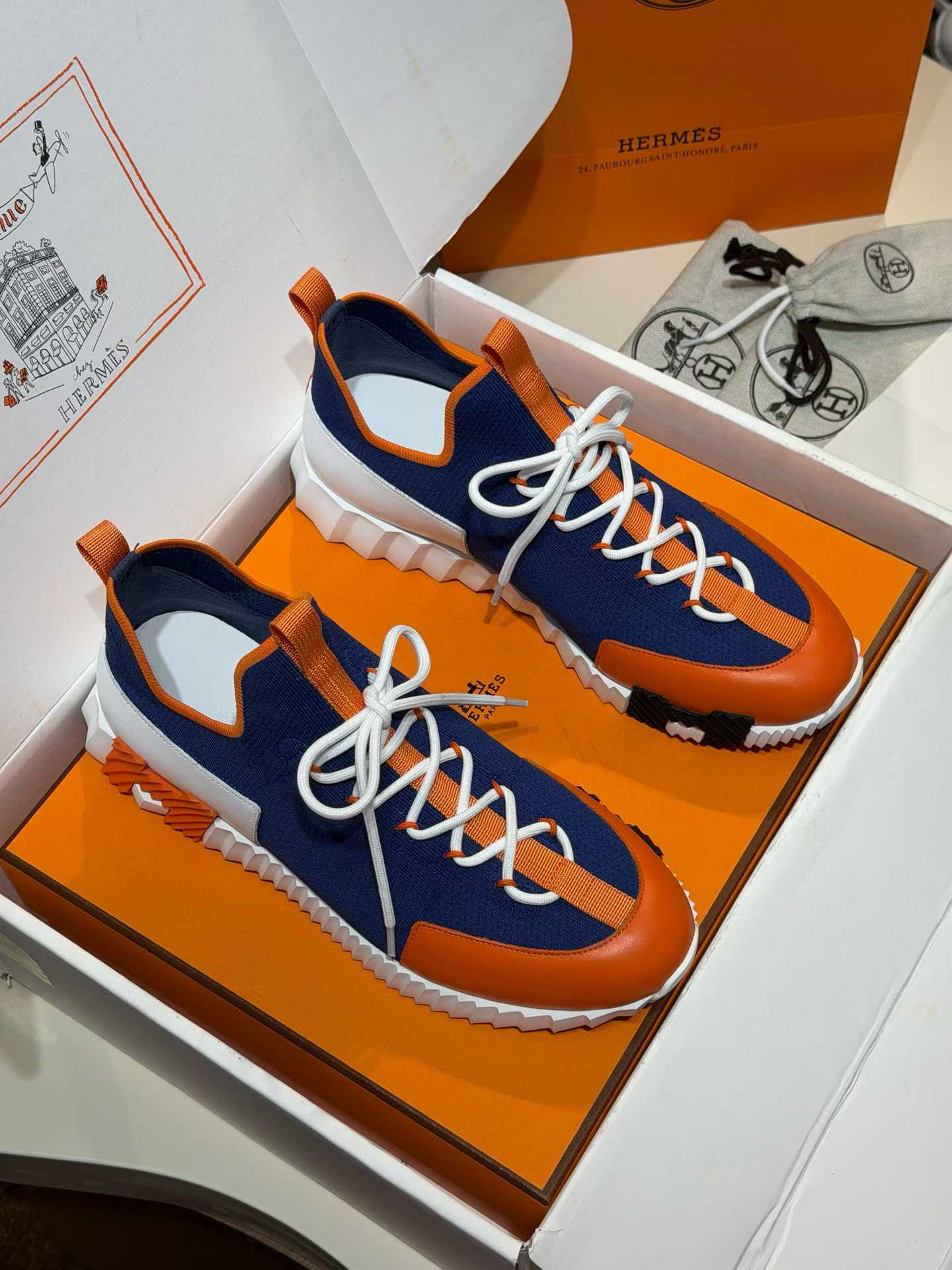 Hermès Lift slip-on sneaker in knit and calfskin with a white rubber sole and orange and black rubber inserts.