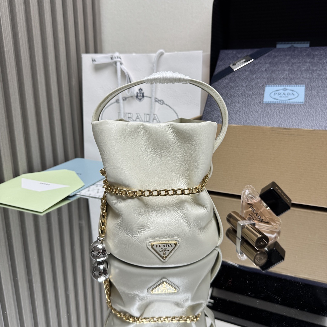 Prada Petit Sac Noir mini bucket bag is crafted from white nappa leather and features a gold-tone chain closure, a matching top handle, and a metallic sphere charm