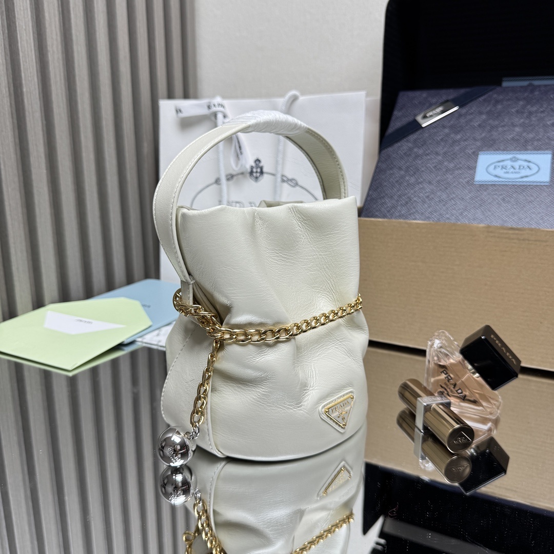 Prada Petit Sac Noir mini bucket bag is crafted from white nappa leather and features a gold-tone chain closure, a matching top handle, and a metallic sphere charm