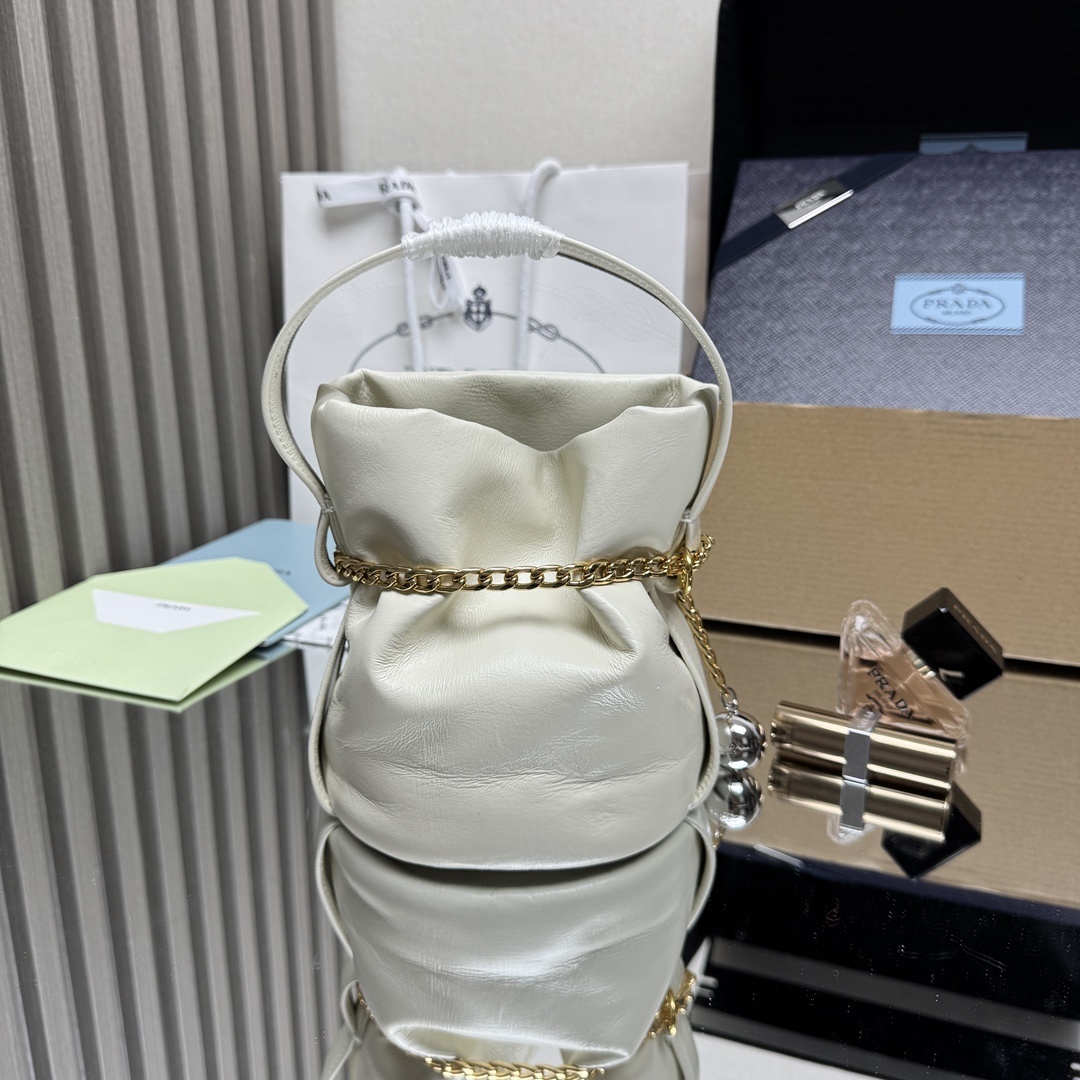 Prada Petit Sac Noir mini bucket bag is crafted from white nappa leather and features a gold-tone chain closure, a matching top handle, and a metallic sphere charm